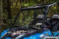 A blue off-road vehicle features a Polaris RZR XP 1000 Full Windshield and “RZR” branded headrests. Parked outdoors among trees, the clear two-piece windshield stands out in the sunlight against the greenery.