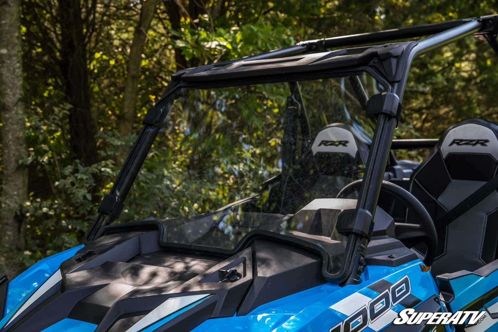 A blue off-road vehicle features a Polaris RZR XP 1000 Full Windshield and “RZR” branded headrests. Parked outdoors among trees, the clear two-piece windshield stands out in the sunlight against the greenery.