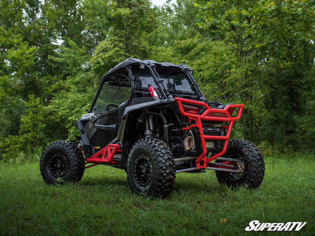 A Polaris RZR XP 1000 with a 3 Long Travel Kit Chromoly Tubed, featuring large rugged tires and a roll cage, is parked on grass in a wooded area. The SuperATV logo appears in the bottom right corner with trees filling the background.