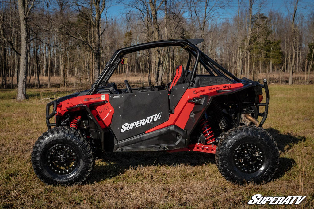 A Polaris RZR XP Turbo with SuperATV branding and protective roll bars, equipped with large rugged tires and Polaris RZR XP Turbo Fender Flares, is parked on dry grass in front of leafless trees under a blue sky.