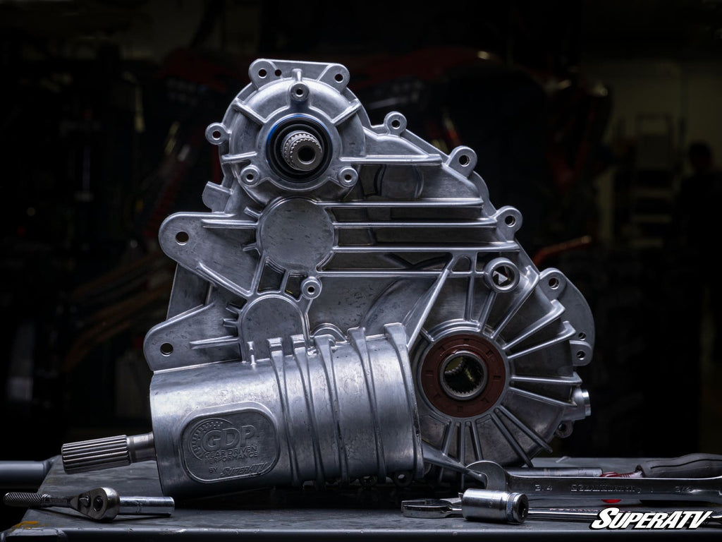 The Polaris RZR Trail S 900 Complete Heavy-Duty Transmission, featuring a silver aluminum case with visible gears and bolts, stands vertically on a dark workbench with tools and a metal pipe in front. The SuperATV logo appears at the bottom right over a d