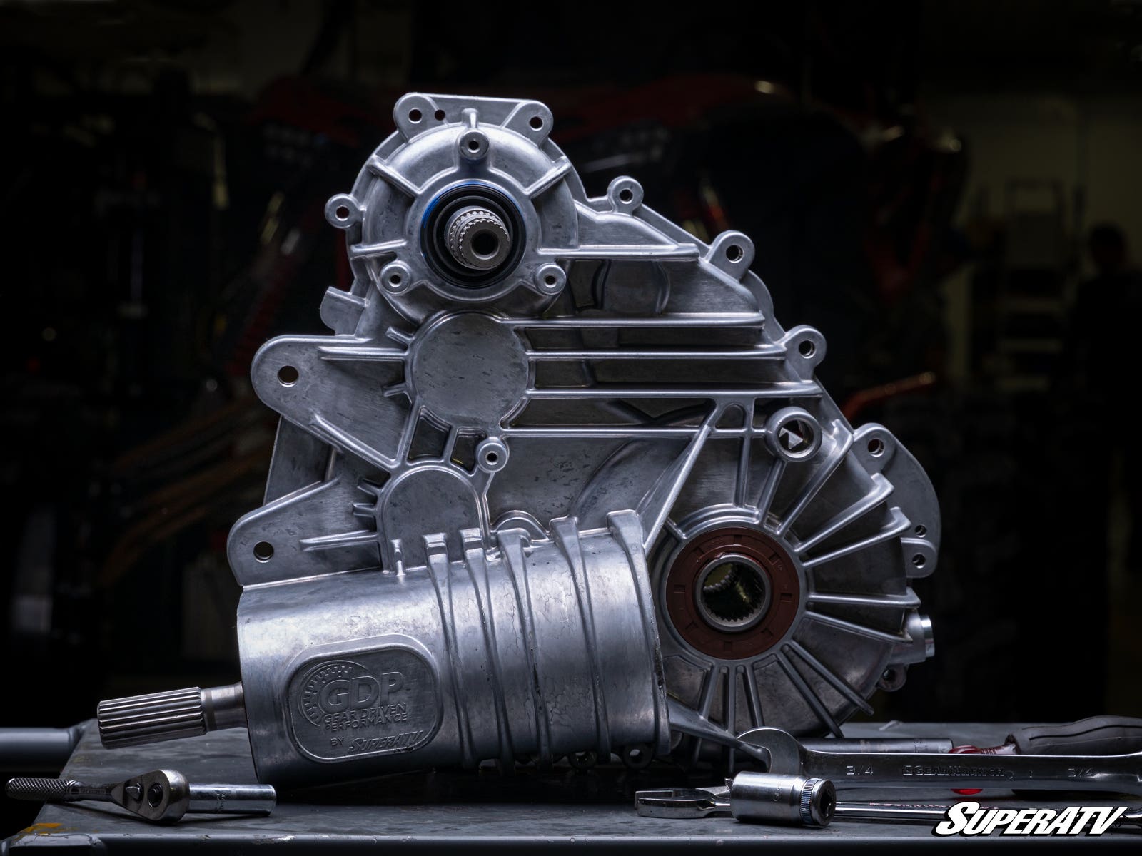 A close-up of a Polaris RZR Trail 900 Complete Heavy-Duty Transmission sits on a workbench with tools. Intricate gears and bolts are visible, set against a dark, blurred background with the SuperATV logo in the corner.