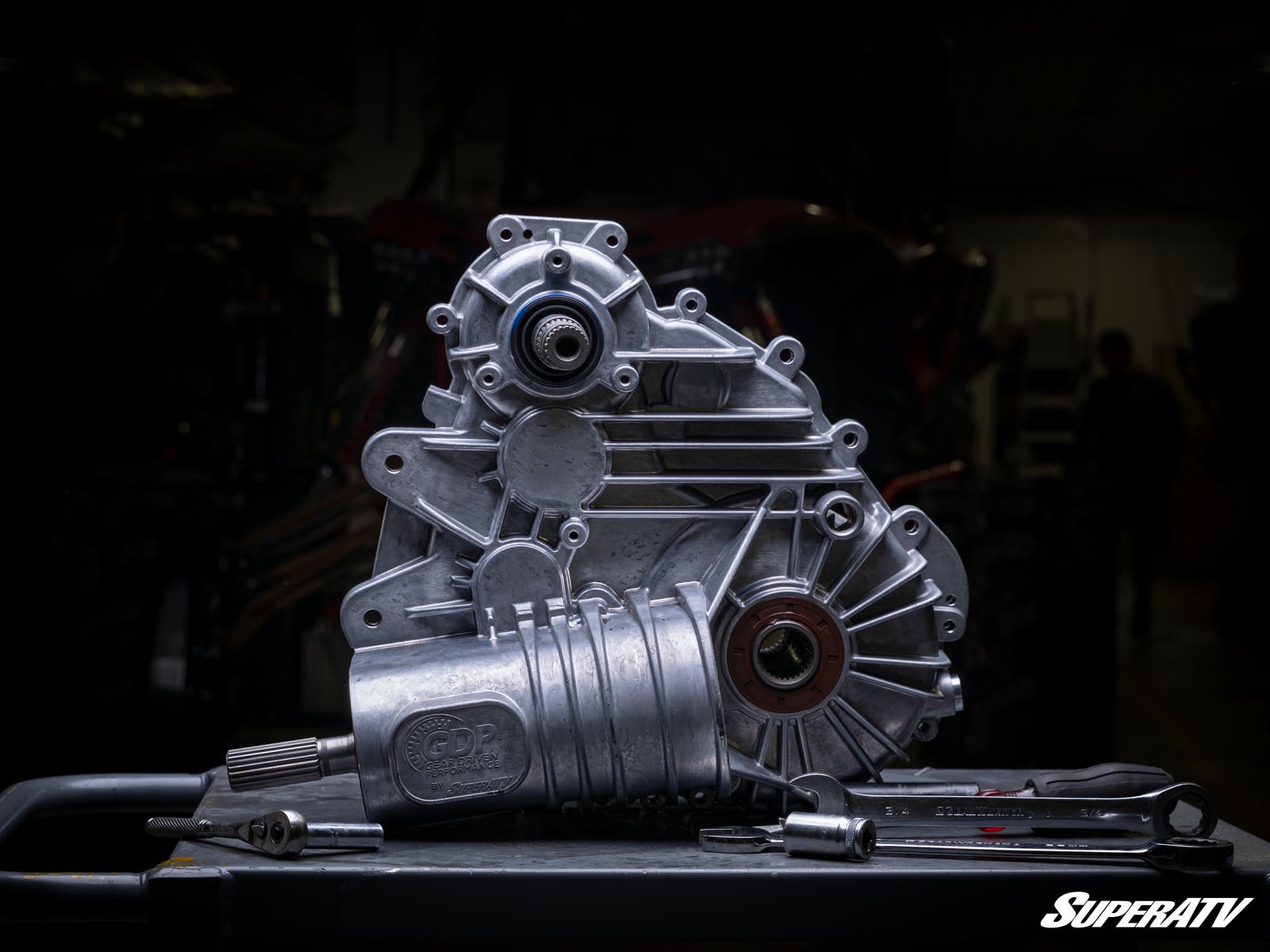 A Polaris RZR XP 1000 Complete Heavy-Duty Transmission with precise machining sits on a dark workbench beside wrenches. The shiny metal assembly stands out against a dim, blurred workshop, branded “SuperATV” in the bottom right corner.