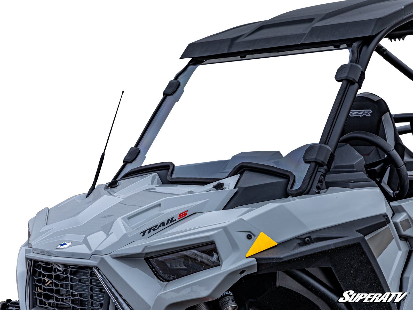 Close-up of the front left side of a gray off-road vehicle equipped with the Polaris RZR Trail 900 Full Windshield, featuring TRAIL S text, tinted glass, angular headlights, side mirror, yellow reflector, and a visible SuperATV logo. Interior is partially