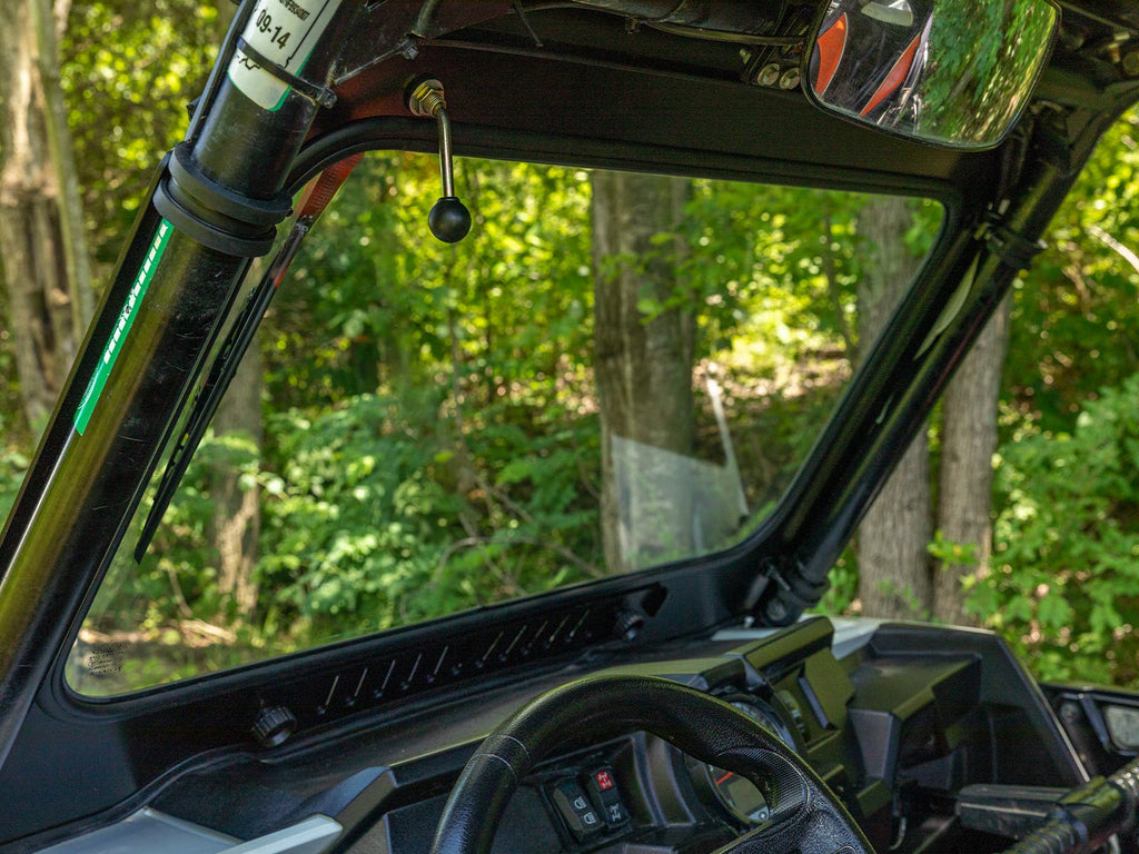 View from inside an off-road vehicle with a Polaris RZR XP Turbo Glass Windshield, showing the steering wheel, dashboard, and roll cage. Sunlight highlights lush forest scenery through the windshield; rearview mirror visible.