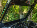 View from inside an off-road vehicle with a Polaris RZR XP Turbo Glass Windshield, showing the steering wheel, dashboard, and roll cage. Sunlight highlights lush forest scenery through the windshield; rearview mirror visible.