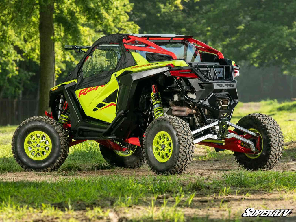 A Polaris RZR Pro R equipped with High-Clearance Billet Radius Arms is parked on grass in a wooded area. The side-by-side ATV features large rugged tires, a protective cage, and the “SuperATV” logo appears in the bottom right corner.