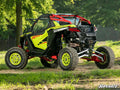 A Polaris RZR Pro R equipped with High-Clearance Billet Radius Arms is parked on grass in a wooded area. The side-by-side ATV features large rugged tires, a protective cage, and the “SuperATV” logo appears in the bottom right corner.