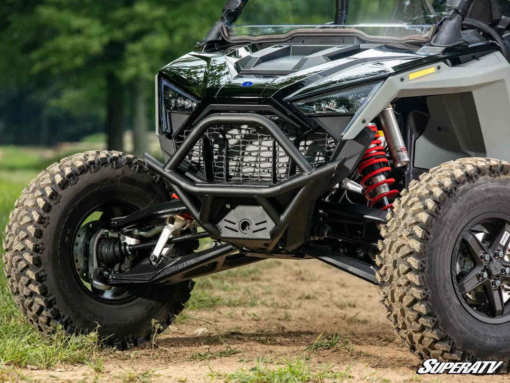 Close-up of a Polaris RZR Pro S Front Bumper with large rugged tires, red coil shocks, and steel brush guard on angular black and gray panels. The SuperATV logo appears at the bottom right against a dirt ground and blurred green trees.
