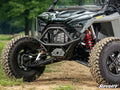 Close-up of the Polaris RZR Turbo R Front Bumper with red shock absorbers, rugged tires, and a SuperATV logo on screen. The scene features a black protective bumper against a backdrop of greenery and dirt.