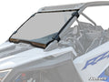A Polaris RZR PRO XP Scratch Resistant Full Windshield with black clamps is mounted on a white UTV, attaching to the roll cage. The clear windshield has a tinted top strip and reveals the vehicle’s interior and steering wheel.