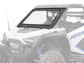 A white off-road vehicle features a Polaris RZR PRO XP Glass Windshield with a black frame, visible bolts, and tilt-out hinge. The vehicle has rugged tires, blue suspension springs, and visible interior seating.
