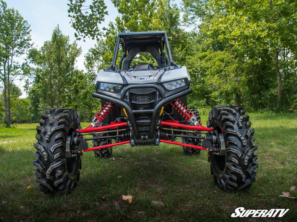 A Polaris RZR RS1 equipped with a 6 Portal Gear Lift and large, deep-tread tires stands on grass facing the camera. Red suspension arms are visible, green trees fill the background, and the SuperATV logo appears in the bottom right corner.