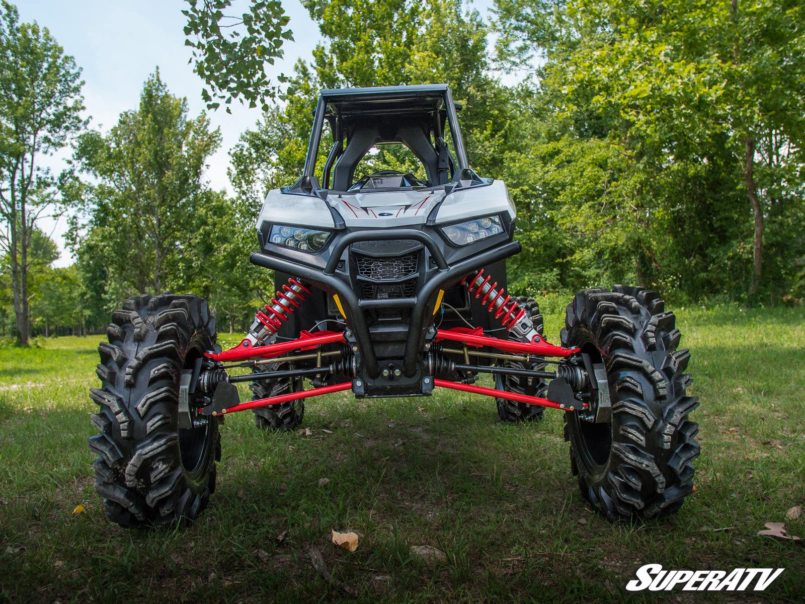 A Polaris RZR RS1 equipped with a 6 Portal Gear Lift and large, deep-tread tires stands on grass facing the camera. Red suspension arms are visible, green trees fill the background, and the SuperATV logo appears in the bottom right corner.