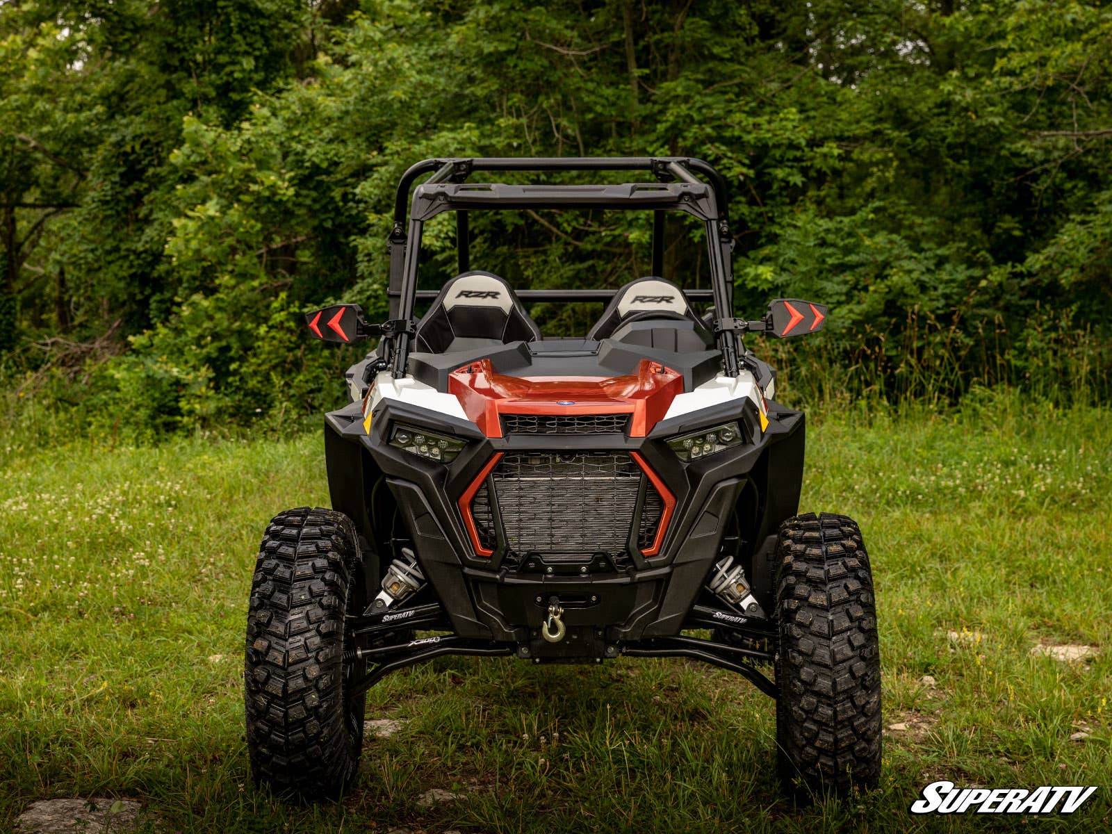 A red and black off-road vehicle featuring Polaris RZR XP Turbo High Clearance 2 Forward Offset A-Arms is parked on grass with thick green trees behind it. It has rugged tires, LED headlights, a roll cage, and SuperATV in white at the bottom right.