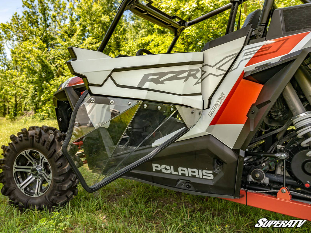 A close-up shows an open side door on a Polaris RZR with a white, orange, and black design and RZR XP and POLARIS logos. The Polaris RZR S4 900 Clear Lower Doors feature a transparent lower panel. Green trees and grass are visible in the background.