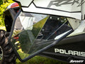 Close-up of Polaris RZR XP 4 1000 Clear Lower Doors, showcasing their angular polycarbonate design with black trim and bolts. The SuperATV logo appears bottom right. Greenery and part of a large rugged rear tire are visible in the background.