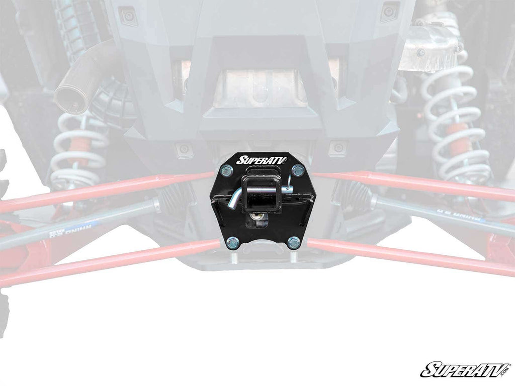 A close-up of a black Polaris RZR RS1 Rear Receiver Hitch mounted on the rear of an off-road vehicle, centered between red suspension arms and silver shocks. The background is faded, with the SuperATV logo in the bottom right corner.