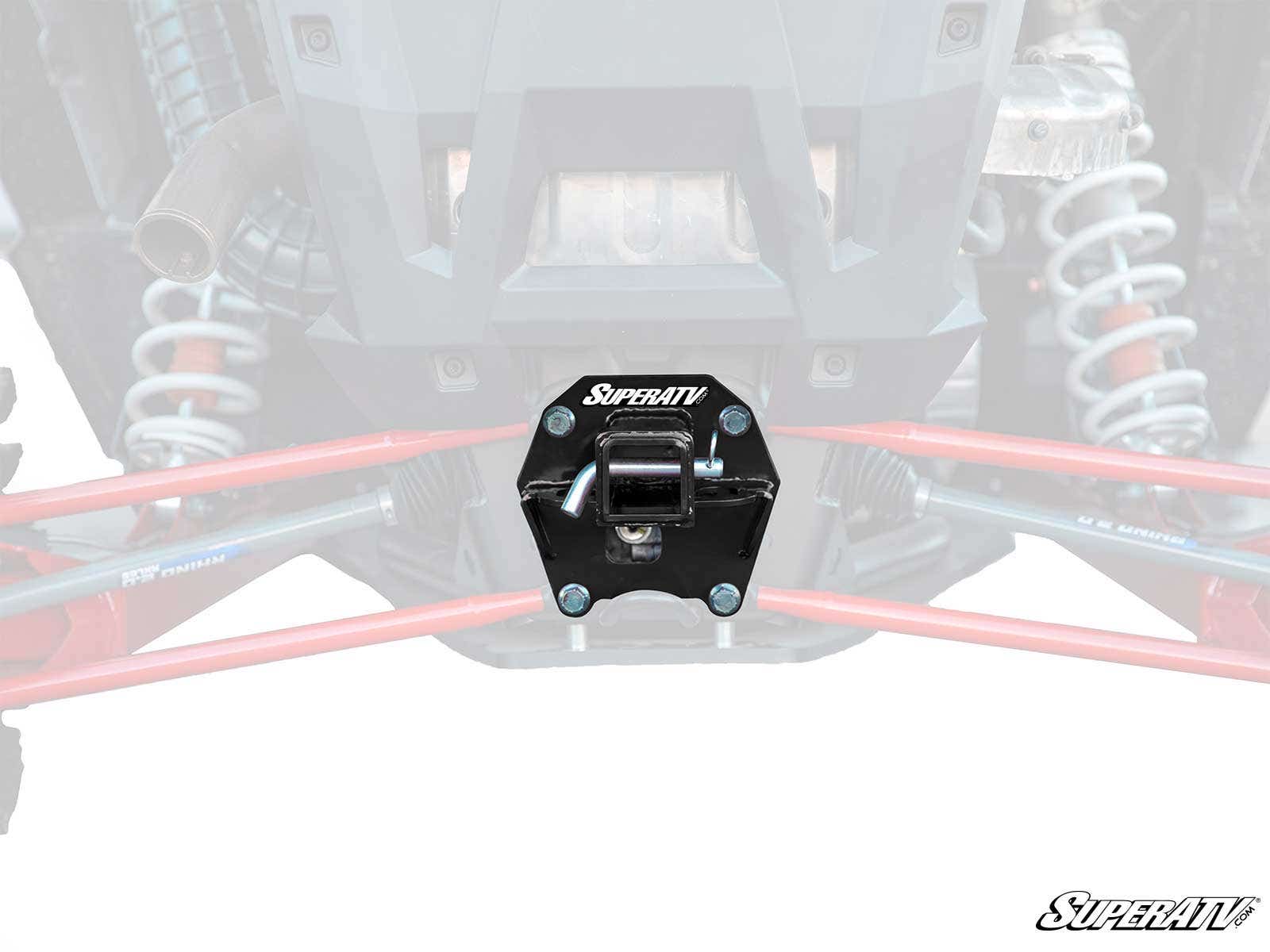 A close-up of a black Polaris RZR RS1 Rear Receiver Hitch mounted on the rear of an off-road vehicle, centered between red suspension arms and silver shocks. The background is faded, with the SuperATV logo in the bottom right corner.