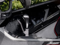 A close-up of the Polaris RZR PRO XP Pistol Grip Gated Shifter with a metal handle inside a vehicle featuring a plain dark interior, red-trimmed seat, SuperATV logo in the corner, and blurred greenery visible through the window.