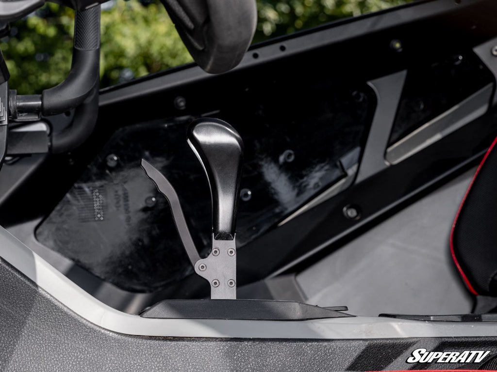 Close-up of a Polaris RZR Turbo R Pistol Grip Gated Shifter with a matte metal base inside a vehicle cockpit. A steering wheel is partially seen on the left, with interior side panel and red-trimmed black seat in the background. SuperATV logo shown.