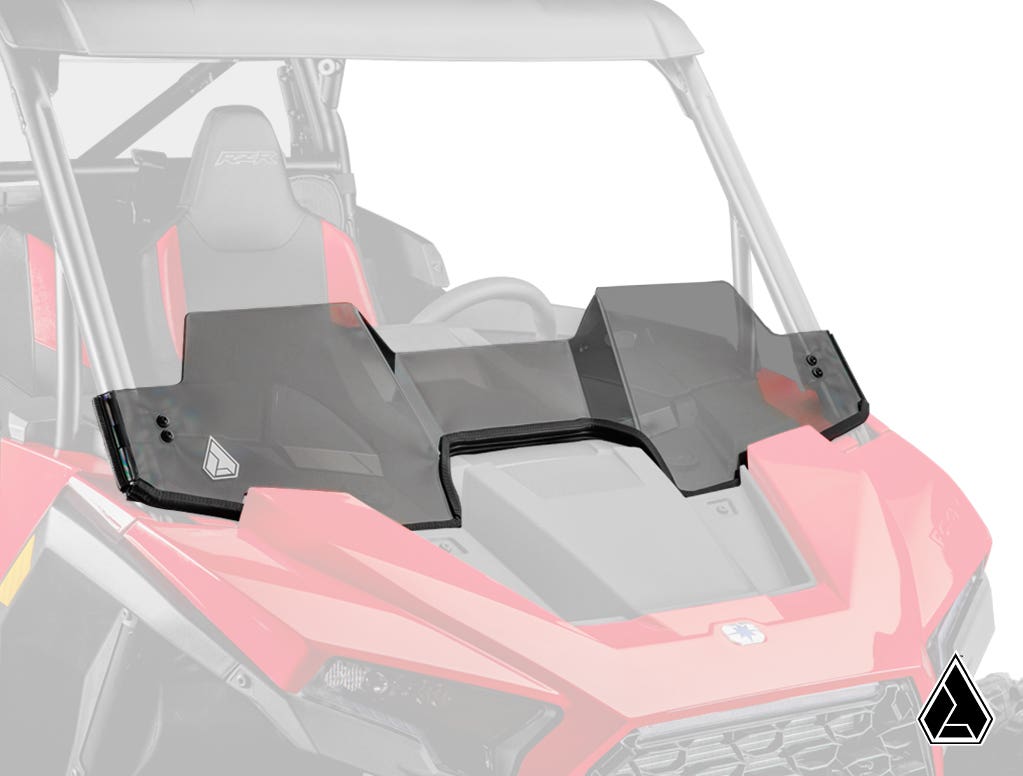 A contoured, tinted Assault Industries Polaris RZR XP Half Windshield is mounted on a red off-road vehicle, featuring two logo decals. The seats, dashboard, steering wheel, and a triangular logo are also visible in the image.
