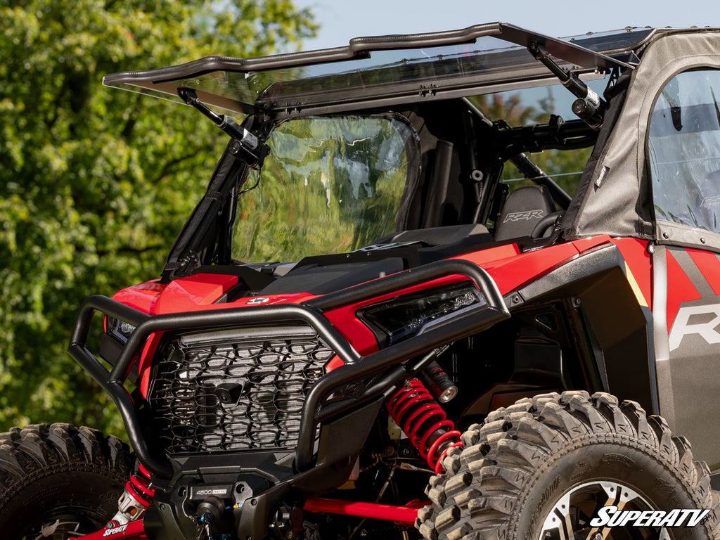 A close-up of a red and black off-road vehicle with large tires features a Polaris RZR XP MaxDrive Power Flip Windshield, partially flipped up. The vehicle is parked outdoors among green trees. SuperATV branding appears in the bottom right corner.
