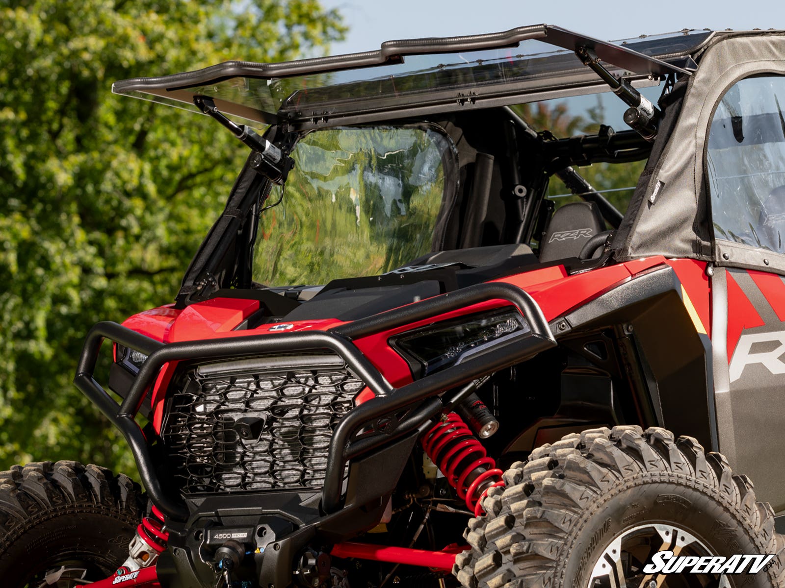 A close-up of a red and black off-road vehicle with large tires features a Polaris RZR XP MaxDrive Power Flip Windshield, partially flipped up. The vehicle is parked outdoors among green trees. SuperATV branding appears in the bottom right corner.