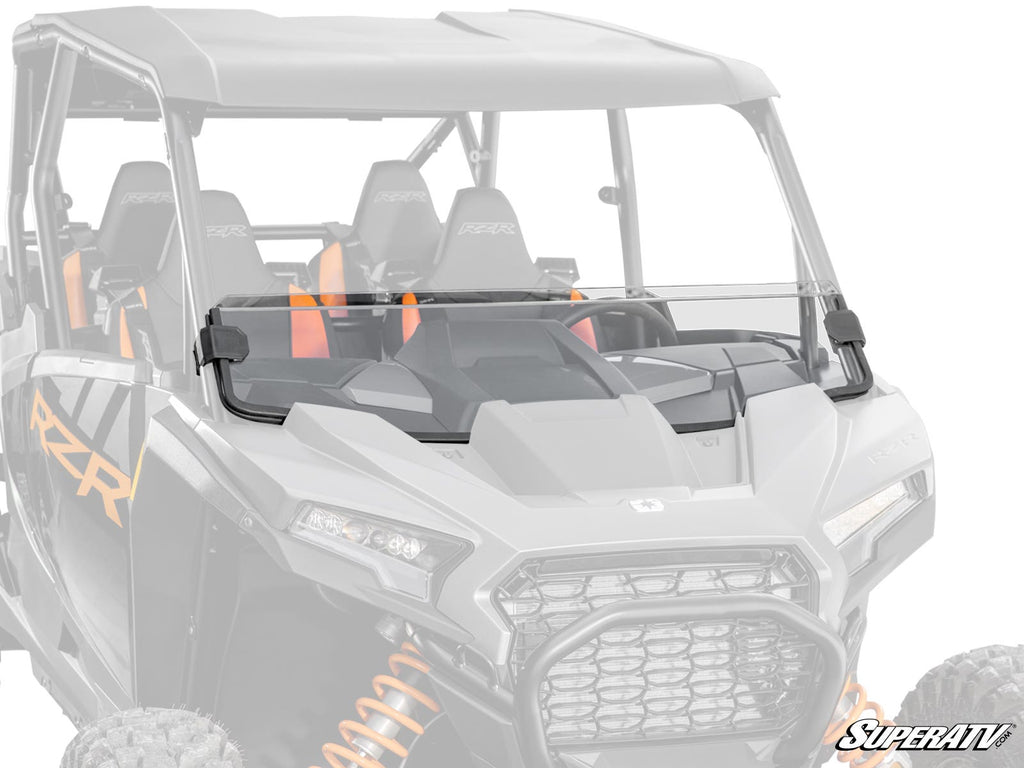 A Polaris RZR XP Half Windshield is mounted on a black side-by-side with orange suspension springs, spanning the front over the dashboard and steering wheel. The SuperATV logo appears in the lower right corner.