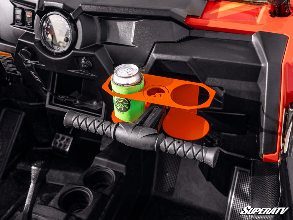 Close-up of a UTV dashboard with a Polaris RZR Grab Bar Cup Holder in bright orange, holding a canned drink in a green sleeve. The interior features a grab bar, foot pedals, gauges, cup holders, and rugged black trim.
