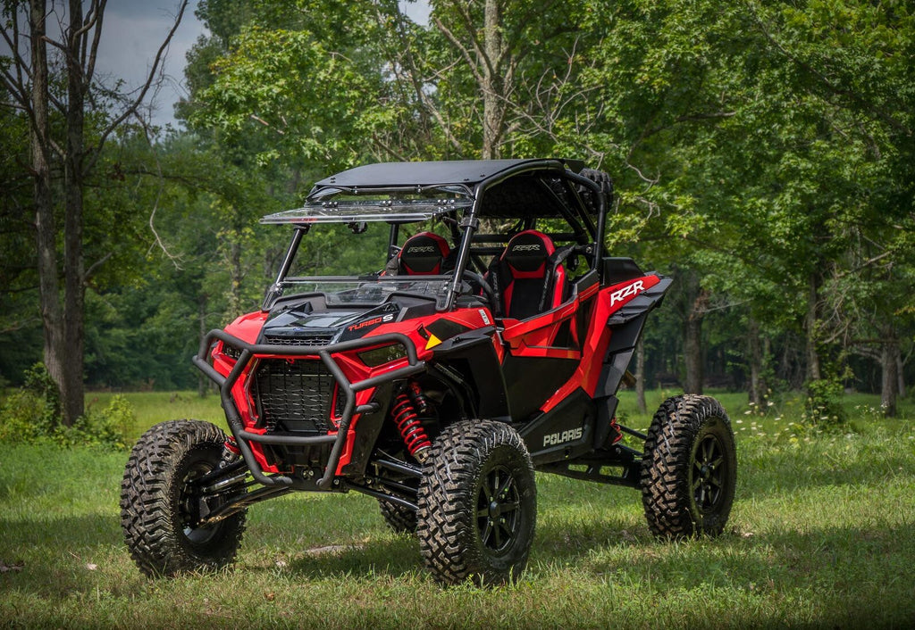 A red and black Polaris RZR featuring the XP Turbo S Flip Windshield is parked on grass in a forest, equipped with rugged tires, roll cage, and two bucket seats under a partly cloudy sky.