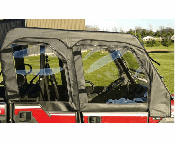 A close-up side view of a Kawasaki Mule Pro-FXT with Soft Upper Doors installed. The black soft doors have clear windows showing the seats and steering wheel. The red utility vehicle is parked on grass near buildings and trees under a clear sky.