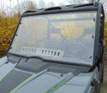 A close-up of an Arctic Cat Prowler Pro with a 1-piece windshield thats clear and slightly reflective, featuring visible wipers. The interior displays two black seats, a steering wheel, and a roll cage. Yellow flowers and bare trees appear behind the vehi