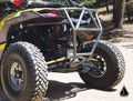 A close-up of Assault Industries High Clearance Radius Rods installed on a Maverick X3 72 model shows the off-road vehicle’s rugged rear and metal bumper on a dirt path among trees, with sunlight and a triangular logo in the lower right corner.