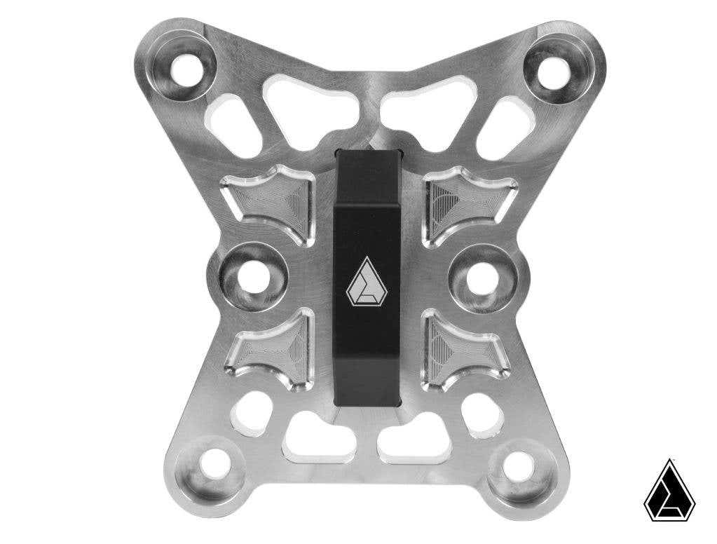 The Assault Industries Billet Radius Rod Chassis Brace for Can-Am Maverick X3 features a CNC-machined silver X-shaped bracket with circular cutouts and a centered black piece displaying a white triangular logo, also shown on the lower right corner.