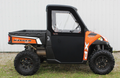 A side view of a Polaris Ranger XP 900 with Full Size Convertible Doors, featuring an orange and black body, black hard cab with clear windows, large rugged tires, and a rear cargo bed, parked on grass by a white metal building.
