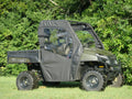 A Polaris Ranger UTV with large off-road tires is parked on grass. It features the Single Cab Soft Door Kit, which has a black fabric enclosure with clear plastic windows. Dense green trees are in the background.