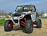 A Polaris Rzr XP 900 with an S3 Power Sports 5 Bracket Lift Kit, featuring large rugged tires, red suspension springs, roll cage, and black accents, is parked on grass with trees and buildings in the background under a blue sky.