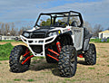 A Polaris Rzr XP 900 with an S3 Power Sports 5 Bracket Lift Kit, featuring large rugged tires, red suspension springs, roll cage, and black accents, is parked on grass with trees and buildings in the background under a blue sky.