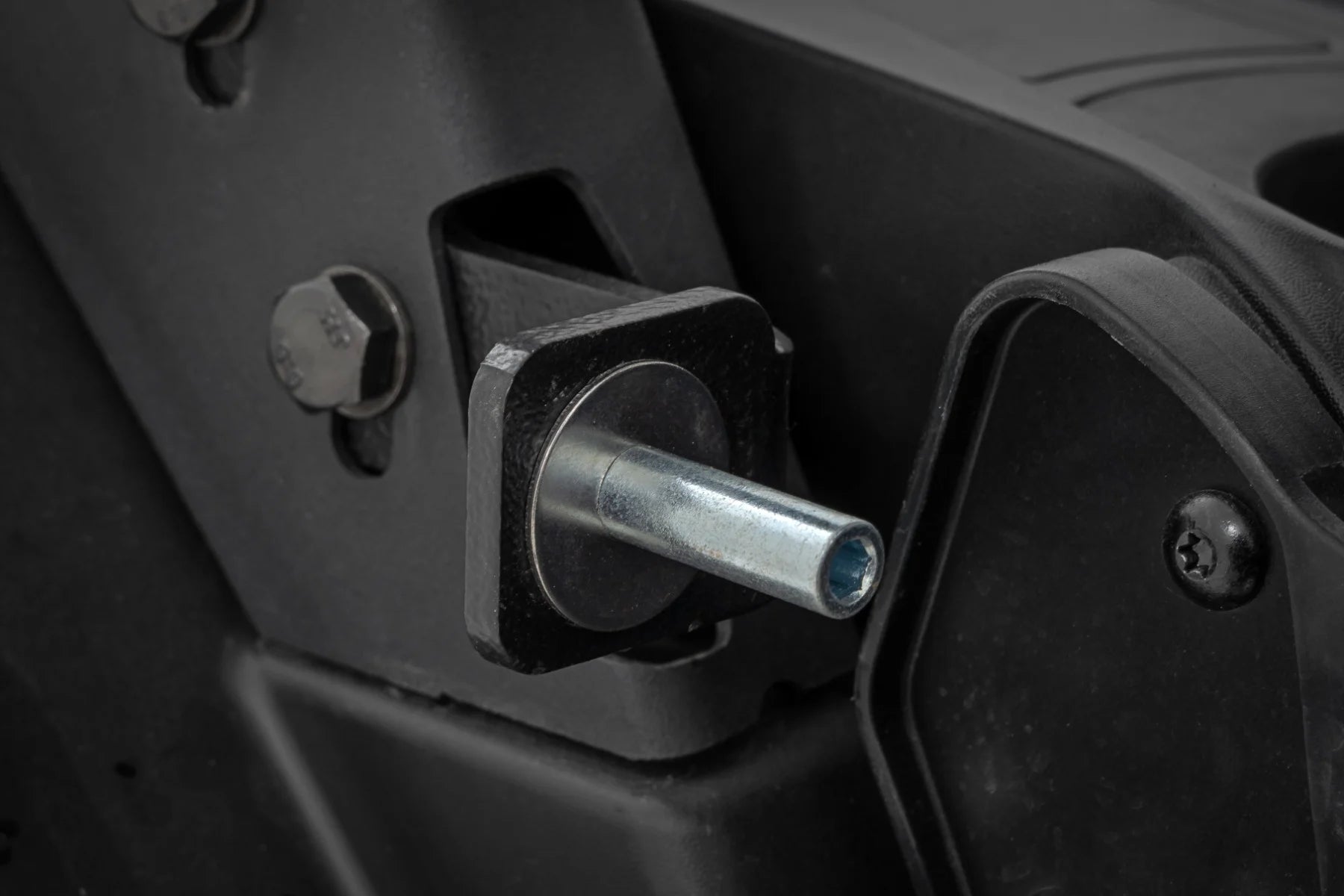 Close-up of a metal pin on a square black bracket attached to a Can-Am Defender 2 Seater Door (HD 5/7/8/9/10). A large hex bolt is nearby, with part of another molded component visible on the right.