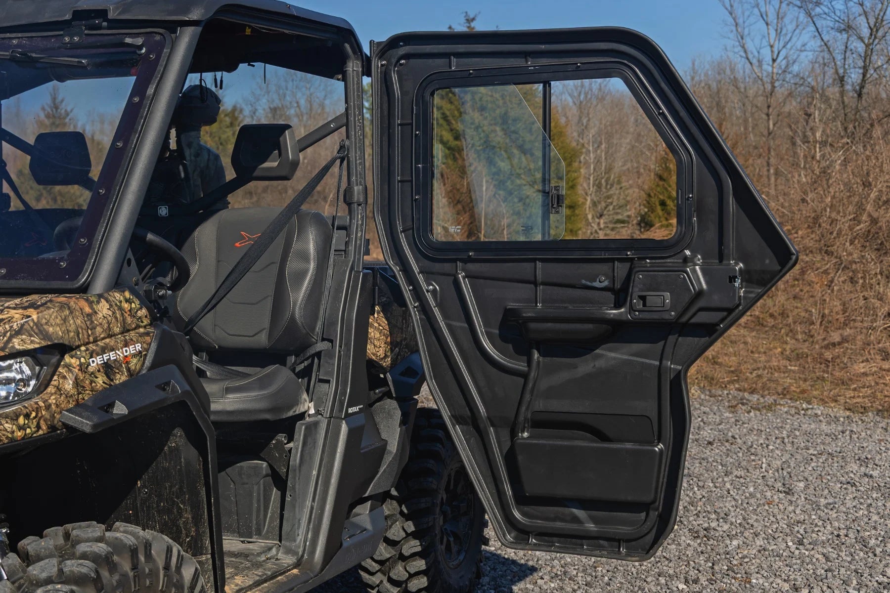 A 2 Seater Can-Am Defender with its driver-side door open is parked on gravel. The black seat and steering wheel are visible inside. Leafless trees and a clear sky in the background suggest late autumn or winter.