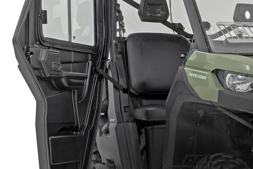A close-up shows the open door of a 2 Seater Can-Am Defender HD 5/HD 7/HD 8/HD 9/HD 10, revealing the black interior with a plain seat, seatbelt, and dashboard. “DEFENDER” is printed on the vehicle’s side.