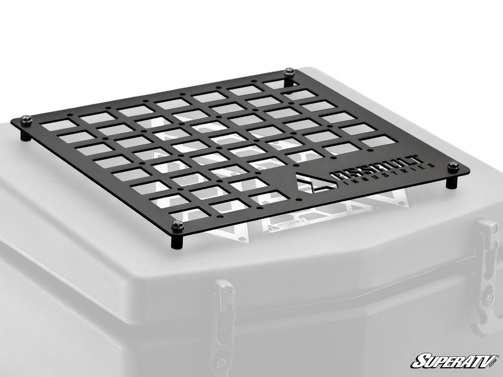 The Assault Industries Cooler/Cargo Box MOLLE Panel is a black metal grid with square openings, mounted atop a gray box. It features the ASSAULT INDUSTRIES logo cut into it and SuperATV branding in the lower right corner, secured by four corner bolts.