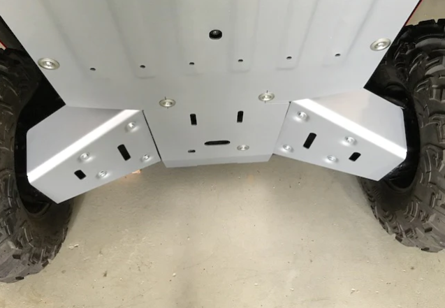 Close-up of Honda Pioneer 1000 Alloy Rear A Arm Guards (2022+) installed beneath the vehicle, showing clean, silver metal guards securely bolted to the frame between large tires, with protective angles and cutouts for ventilation or mounting.