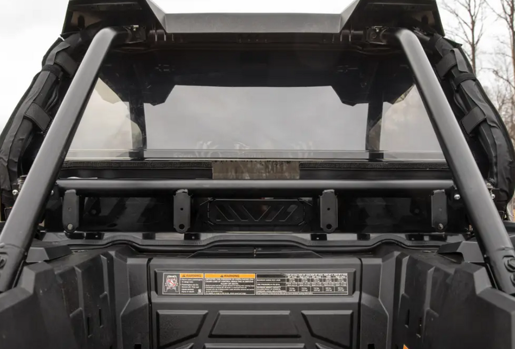 Close-up of the Polaris Rzr Xp 1000 Rear Vented Windshield installed, showing the rear cargo area, roll cage, and visible safety labels inside. The transparent windshield reveals blurred leafless trees in the background.