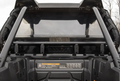 Close-up of the Polaris Rzr Xp 1000 Rear Vented Windshield installed, showing the rear cargo area, roll cage, and visible safety labels inside. The transparent windshield reveals blurred leafless trees in the background.