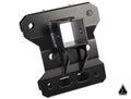 The Assault Industries Heavy Duty Rear Chassis Brace with Tow Hitch for Can-Am Maverick X3 is a black metal bracket featuring a square central opening, multiple screw holes, cutouts, angled edges, raised details, and a triangle logo at the bottom right.