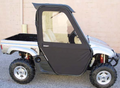 A Yamaha Rhino equipped with Framed Convertible Doors, featuring a black soft door with a clear window, is parked on pavement. The silver off-road utility vehicle has large rugged tires and a small truck bed. Background: beige brick wall.