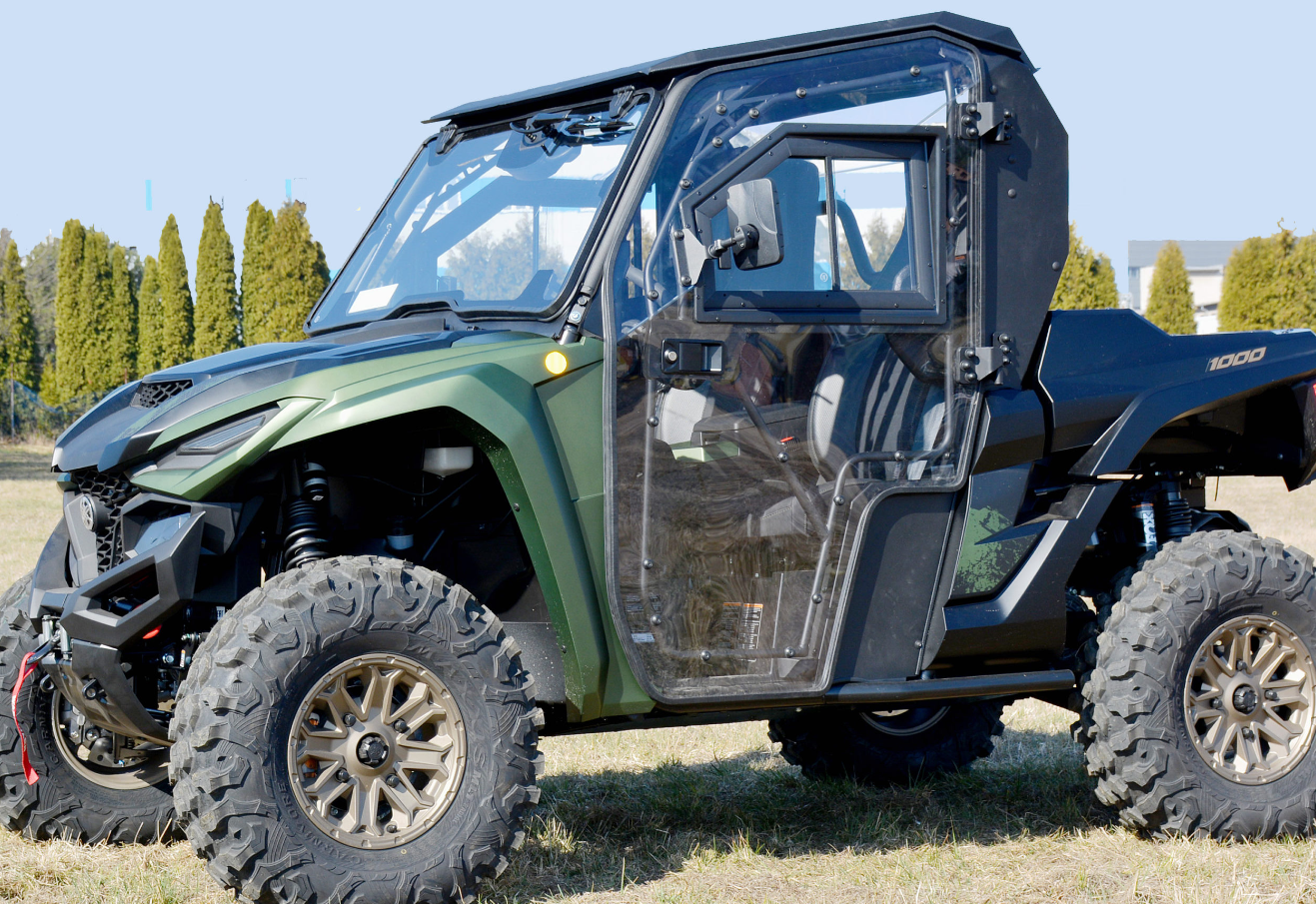 A Yamaha RMAX2 1000 Full Hard Cab in green and black with large rugged tires is parked on grass. Its transparent, open door reveals visible seats and a sturdy frame, with tall evergreens and a blue sky in the background.