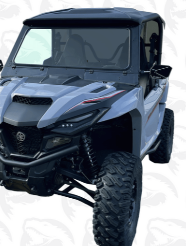 The Yamaha Rmax 1000 Glass Windshield is shown on a rugged, light blue off-road vehicle with large knobby tires, black grill, roof, angular headlights, high ground clearance, and a faint shark-patterned background.